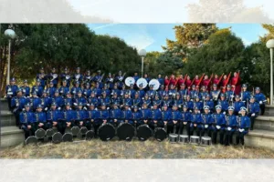 Large marching band in blue uniforms poses on outdoor steps with drums in front of them.
